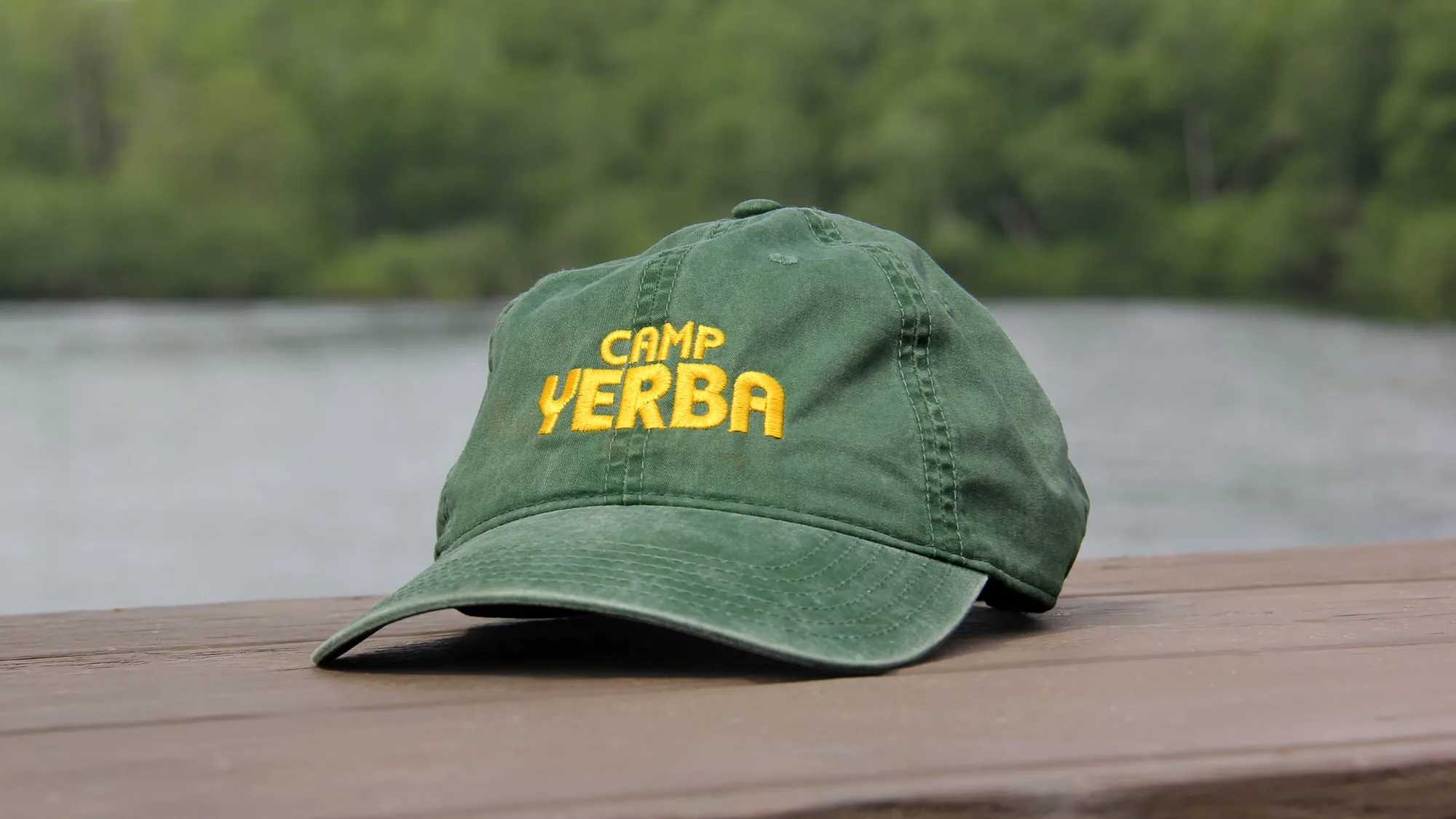 Green Camp Yerba cap with yellow embroidery on a railing by the lake